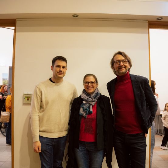 Drei Menschen stehen vor einer weißen Wand. Der Mann auf der linken Seite trägt einen beigen Pullover. Die Frau in der Mitte trägt ein rotes Oberteil, einen Schal und eine Brille. Der Mann auf der rechten Seite trägt ein rotes Rollkragenhemd, eine schwarze Jacke und eine Brille. Im Hintergrund stehen weitere Menschen und ein Fahrrad.