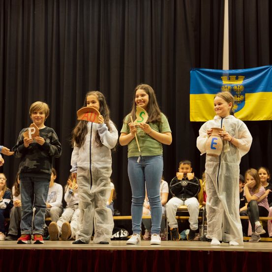 Fünf Kinder in Schutzanzügen stehen auf der Bühne und halten Schilder mit Buchstaben. Dahinter ein schwarzer Hintergrund mit einer ukrainischen Flagge. Andere Kinder sitzen auf Bänken und schauen zu.