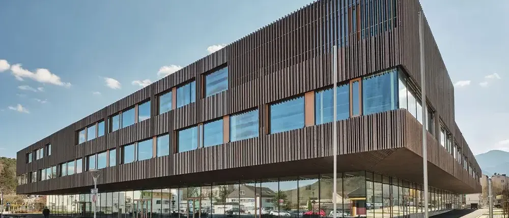 Ein modernes Gebäude mit großen Glasfenstern und einer braunen Außenfassade, die den blauen Himmel und Wolken reflektiert.