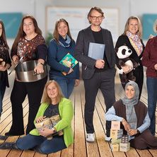 Eine Gruppe von Erwachsenen steht und sitzt auf einem Holzboden und hält verschiedene Gegenstände wie ein Buch, Kuscheltiere und Trommeln. Die Personen lächeln und scheinen für ein Foto zu posieren.