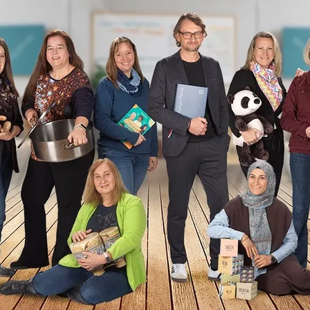 Eine Gruppe von Erwachsenen steht und sitzt auf einem Holzboden und hält verschiedene Gegenstände wie ein Buch, Kuscheltiere und Trommeln. Die Personen lächeln und scheinen für ein Foto zu posieren.