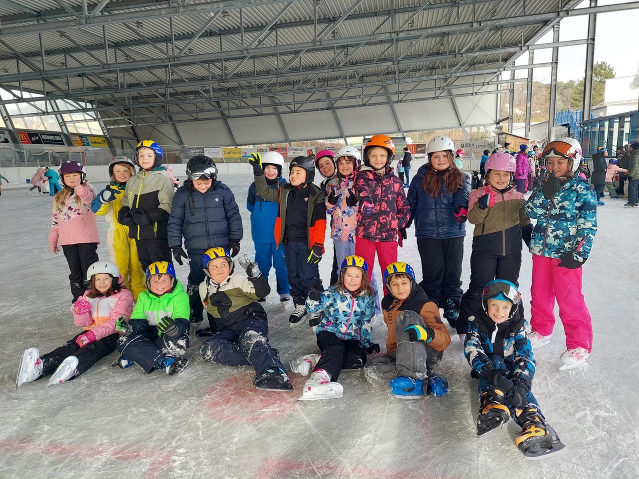 Eine Gruppe von Kindern in Winterkleidung und Helmen posiert für ein Foto auf einer überdachten Eisbahn. Sie lächeln und scheinen sich zu amüsieren.