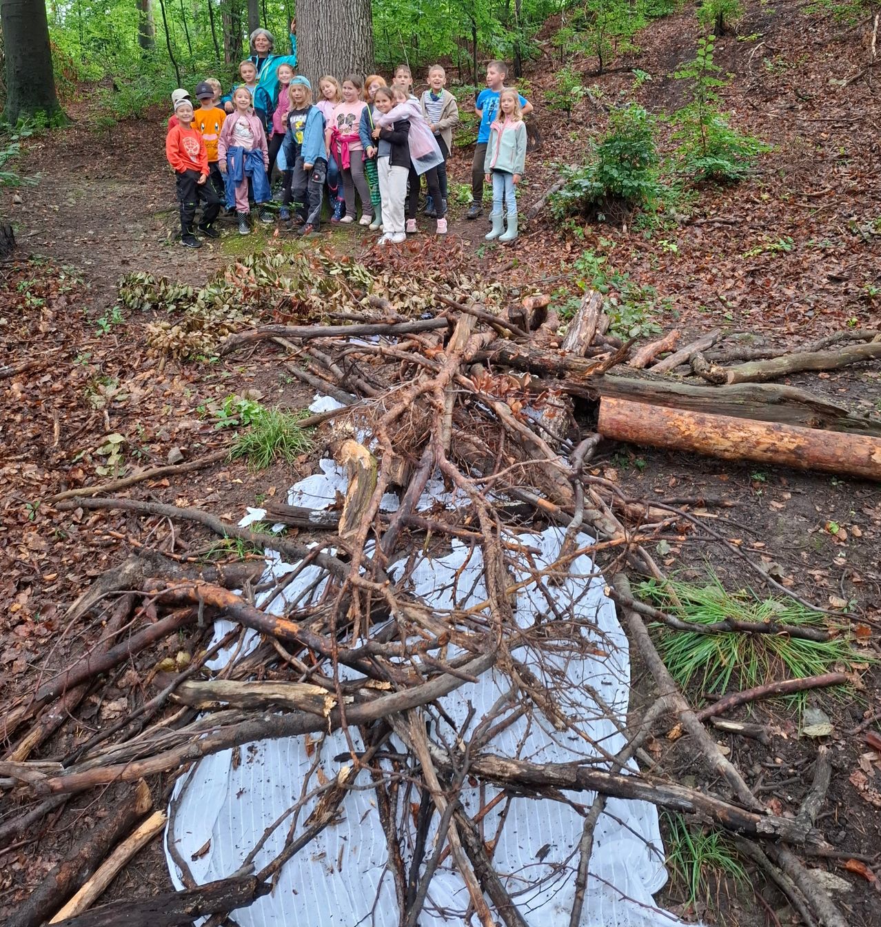 Eine Gruppe von Kindern und Erwachsenen posiert neben einem großen Haufen von Ästen in einem Wald. Sie befinden sich in einer Lichtung mit einem Baumstamm im Hintergrund.