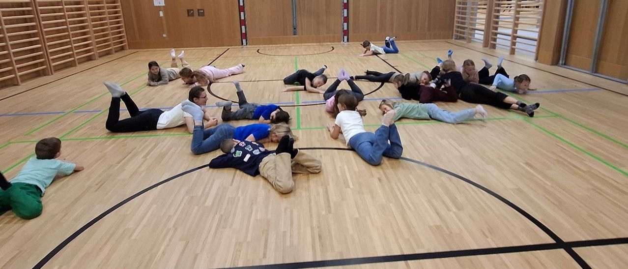 Eine Gruppe von Kindern und Erwachsenen liegt auf dem Hallenboden, wobei einige Erwachsene in der Nähe sind, und bildet einen Kreis mit einem Kind im Inneren.