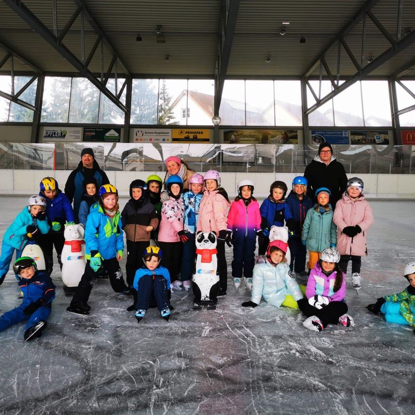 Eine Gruppe von Kindern und Erwachsenen in Winterkleidung und Helmen posiert für ein Foto auf einer überdachten Eisbahn.