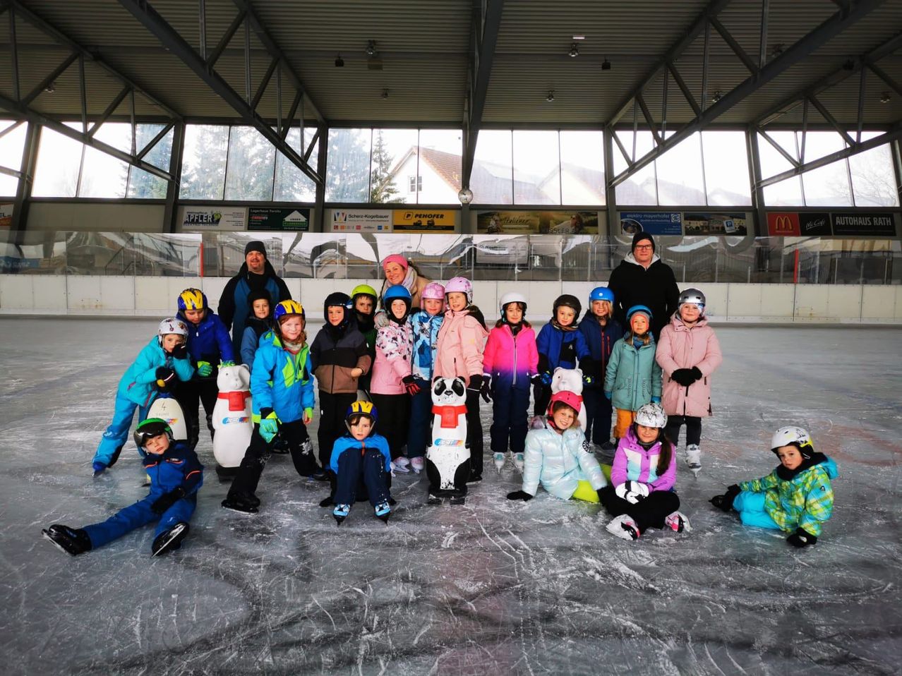 Eine Gruppe von Kindern und Erwachsenen in Winterkleidung und Helmen posiert für ein Foto auf einer überdachten Eisbahn.