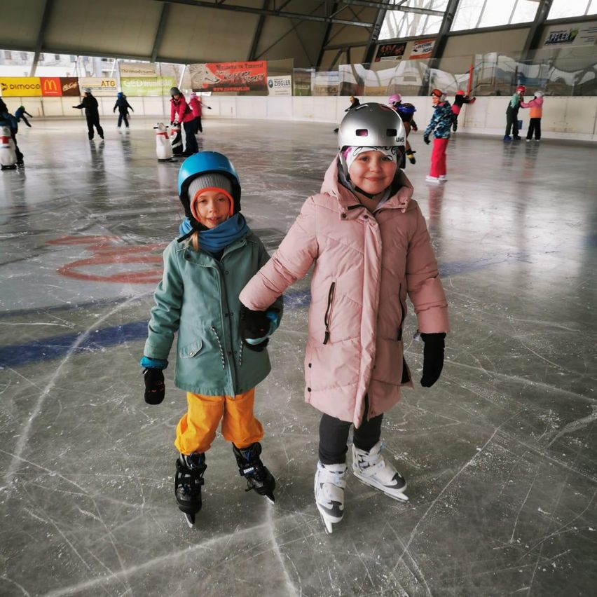 Zwei kleine Kinder, beide mit Helmen und Wintermänteln, skaten gemeinsam auf einer überdachten Eisbahn. Sie halten Händchen. Andere Menschen skaten im Hintergrund.