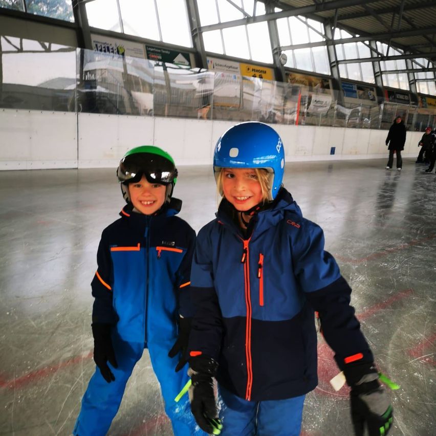 Zwei kleine Kinder, die Helme und Winterkleidung tragen, stehen auf einer überdachten Eisbahn. Sie lächeln und halten Eishockeyschläger in den Händen. Dahinter ist eine Glaswand und eine Person, die geht.