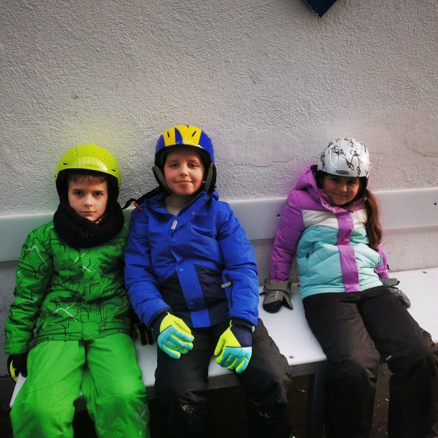 Drei Kinder, gekleidet in bunte Winterkleidung und Helmen, sitzen zusammen auf einer Bank und lächeln für die Kamera.