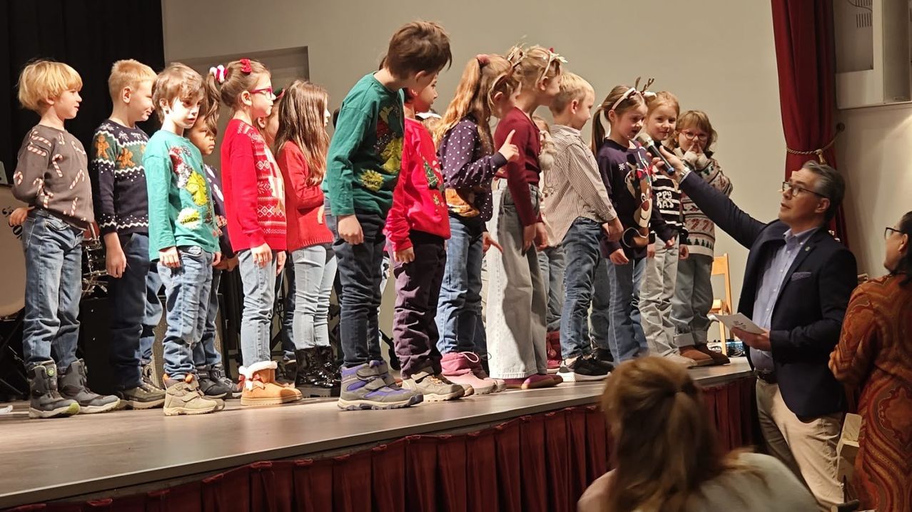 Eine Gruppe von Kindern in festlichen Pullovern steht auf der Bühne und singt mit einem Mikrofon in der Mitte.