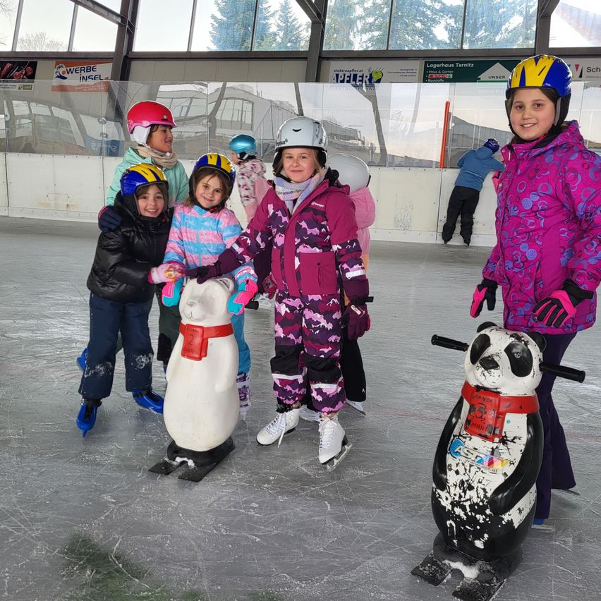 Eine Gruppe junger Kinder mit Helmen und Winterkleidung skatet auf einer Eisbahn. Einige halten sich an einem pinguinförmigen Skulptur fest. Ein anderes Kind skatet im Hintergrund.