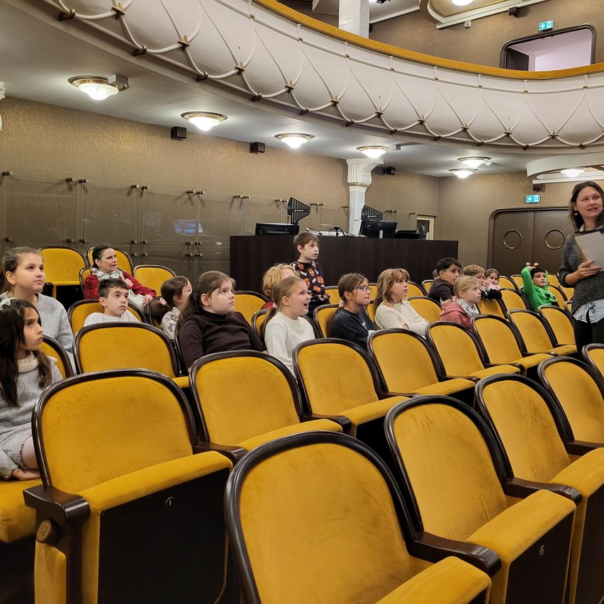 In einem Theater steht eine Frau vor einem Publikum aus Kindern. Das Theater ist mit gelben gepolsterten Sitzen, einem Podium und einer Galerie mit Geländern ausgestattet.