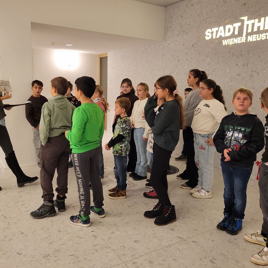 Eine Gruppe von Kindern in verschiedenen Kleidungsstücken steht in einer Reihe in einem Saal. Ein Erwachsener hält vor ihnen ein Papier. Die Wand hinter ihnen hat den Text Stadt Theater Wiener Neust.