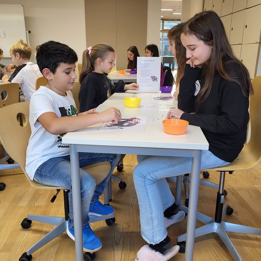 Mehrere Kinder sitzen in einem Klassenzimmer um einen Tisch herum und beschäftigen sich mit einer Aktivität. Ein Junge und ein Mädchen schauen sich etwas auf dem Tisch an. An der Wand befindet sich eine Tafel.