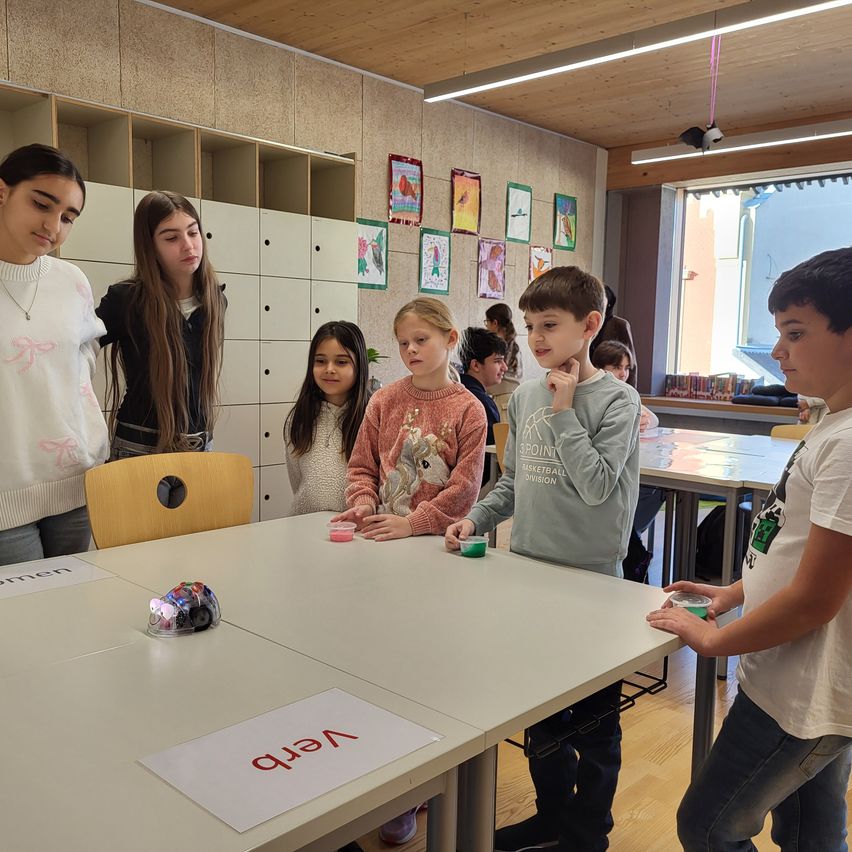 Mehrere Kinder und eine Lehrerin sind um einen Tisch mit einem Roboter versammelt. Auf dem Tisch steht das Wort 'NEAT'. Sie schauen den Roboter an.