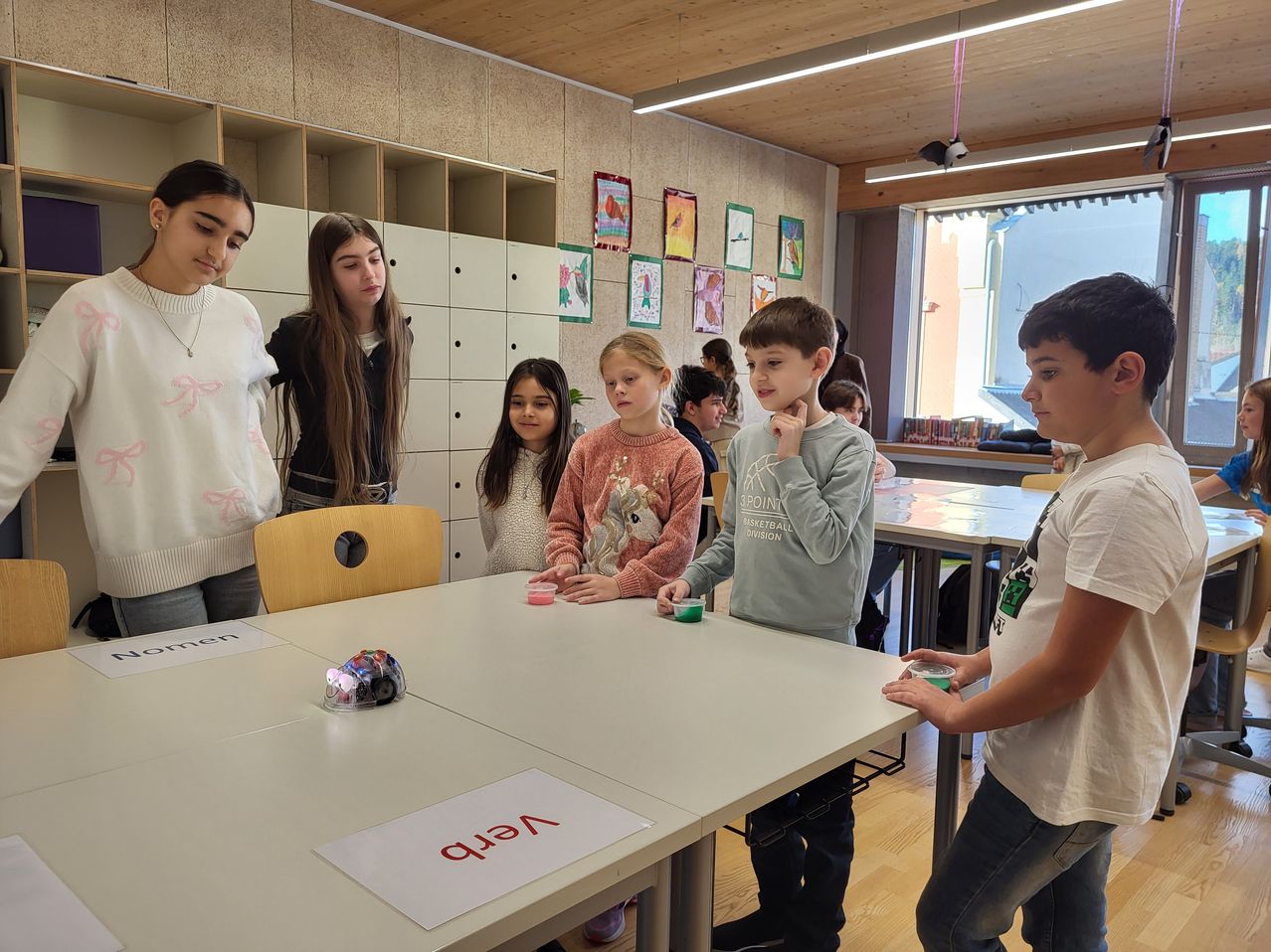 Mehrere Kinder und eine Lehrerin sind um einen Tisch mit einem Roboter versammelt. Auf dem Tisch steht das Wort 'NEAT'. Sie schauen den Roboter an.