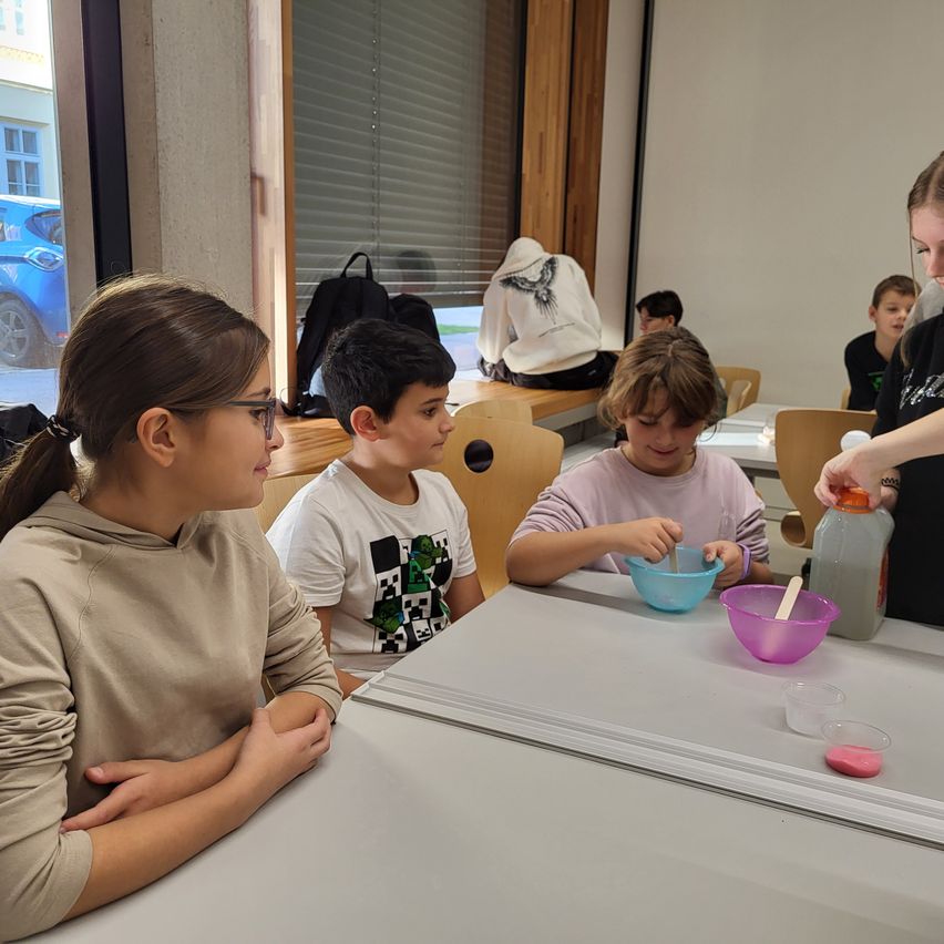In einem Klassenzimmer beobachten drei Kinder, wie eine Frau eine rosa Flüssigkeit in eine Schüssel gießt. Sie sitzen aufmerksam an einem Tisch, mit einer blauen Schüssel und einer Flasche in der Nähe. Hinter ihnen sitzt eine Person auf einer Bank mit einem Rucksack.