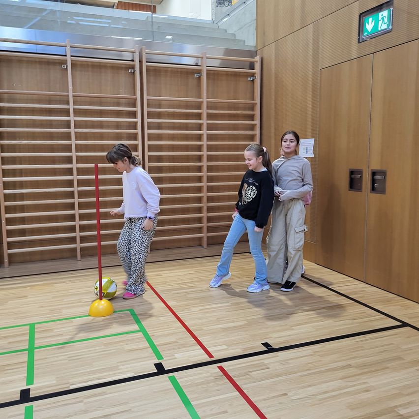 Drei Mädchen spielen ein Spiel mit einem Ball an einem Stock in einer Turnhalle. Sie tragen Freizeitkleidung. Der Boden ist mit Linien markiert.