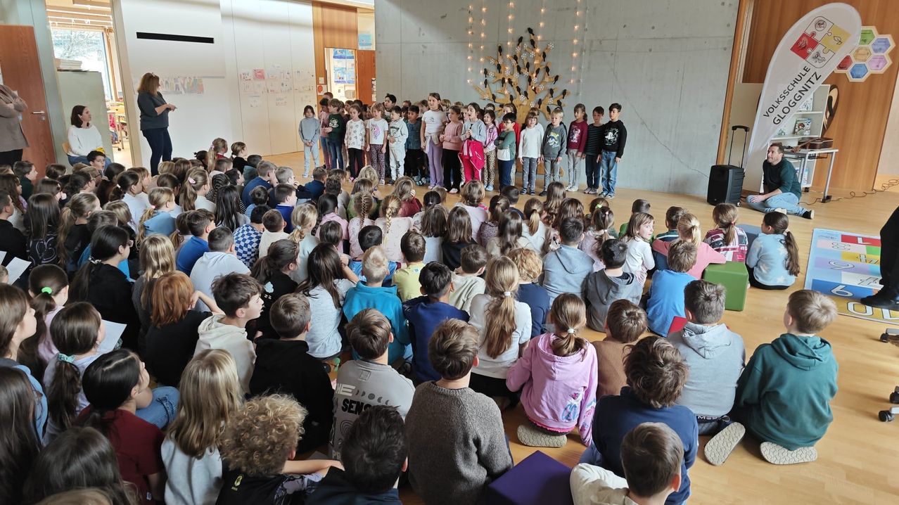 Eine Gruppe von Kindern ist in einem Raum versammelt, einige stehen vorne und andere sitzen auf dem Boden. Die Kinder tragen Freizeitkleidung. Ein dekorativer Baum mit Lichtern ist an der Wand hinter ihnen befestigt.