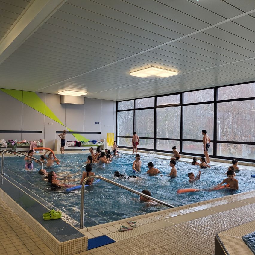 Ein großes Hallenbad, gefüllt mit schwimmenden und spielenden Kindern, beleuchtet durch Deckenleuchten und natürliches Licht aus großen Fenstern.