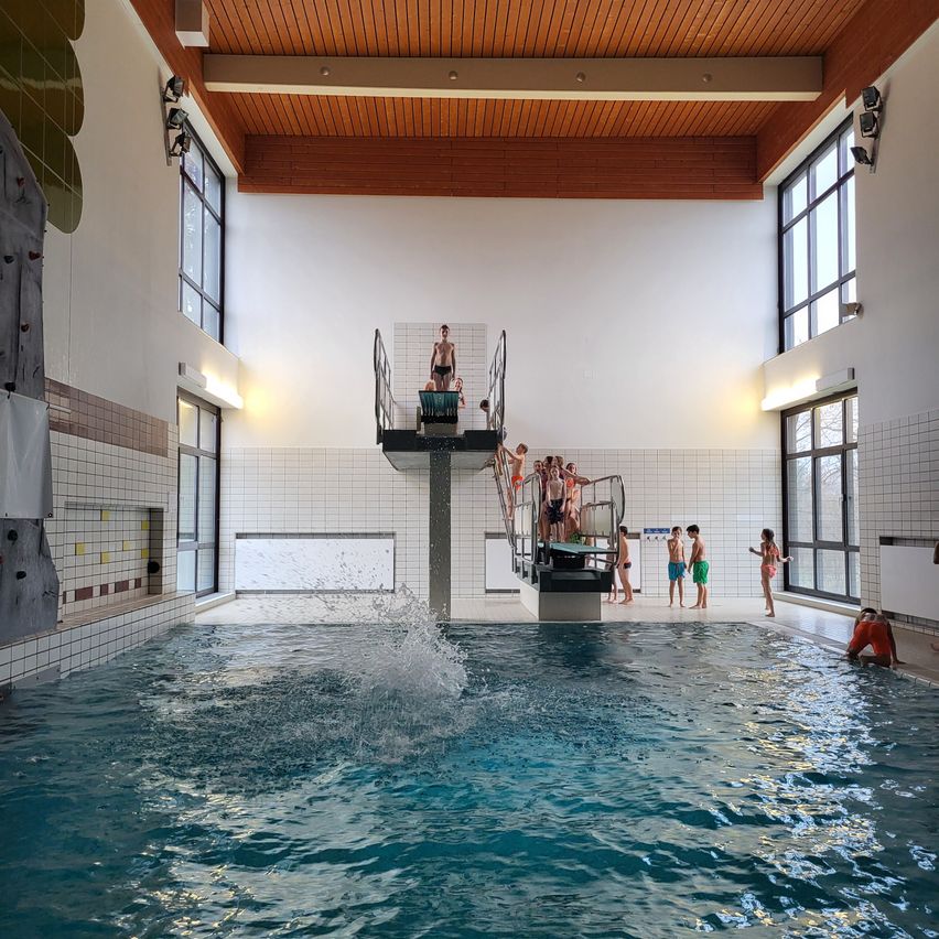 Ein großes Hallenschwimmbad mit mehreren Kindern, die auf der Sprungturm stehen und ein Kind springt ins Wasser. Das Becken ist von weißen Fliesenwänden umgeben und hat eine Holzdecke.
