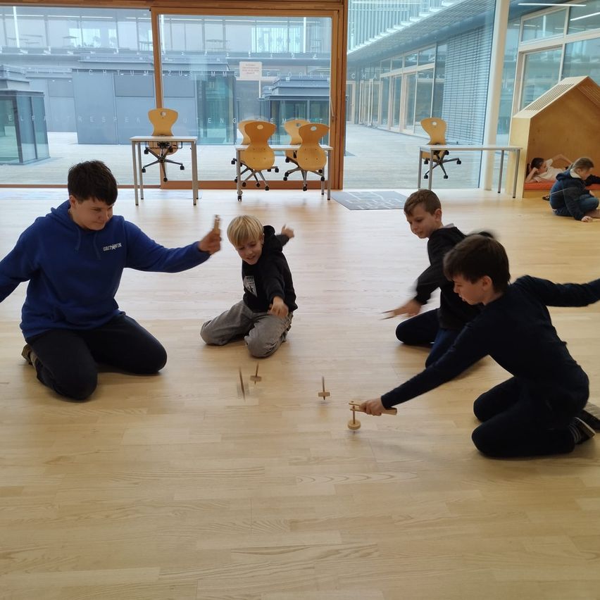 Mehrere Kinder spielen mit Holzspielzeug auf einem Holzboden in einem Raum mit Glaswänden. Ein Junge in einer blauen Hoodie greift nach einem Jungen in einer schwarzen Jacke.