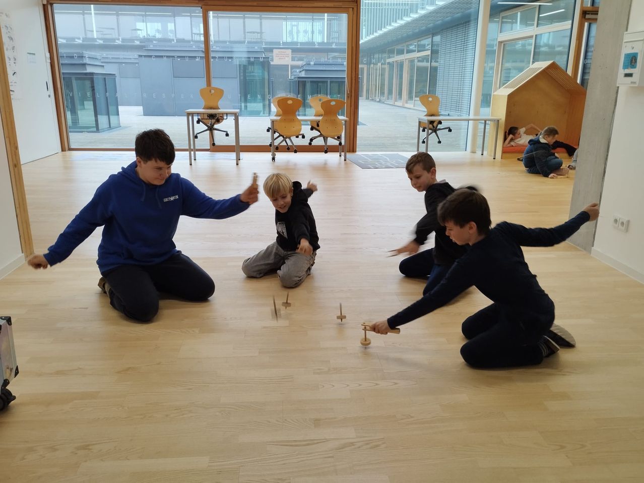Mehrere Kinder spielen mit Holzspielzeug auf einem Holzboden in einem Raum mit Glaswänden. Ein Junge in einer blauen Hoodie greift nach einem Jungen in einer schwarzen Jacke.