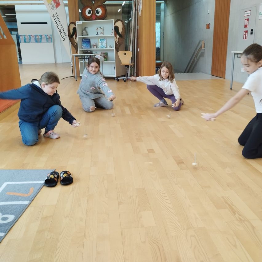 Vier Kinder spielen ein Spiel auf einem Holzboden. Jedes Kind hält einen kleinen Stock, und ein Kind greift nach dem Stock eines anderen. Sie tragen Freizeitkleidung. Hinter ihnen steht ein Bücherregal und ein Schreibtisch.