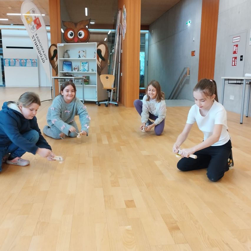 Vier Mädchen spielen auf einem Holzboden, vermutlich in einer Bibliothek. Jede hält ein kleines Objekt, wahrscheinlich ein Spielstein. Sie tragen Freizeitkleidung und scheinen konzentriert zu sein. Hinter ihnen sind eine Eule-Statue, ein Regal mit Büchern und ein Tisch zu sehen.