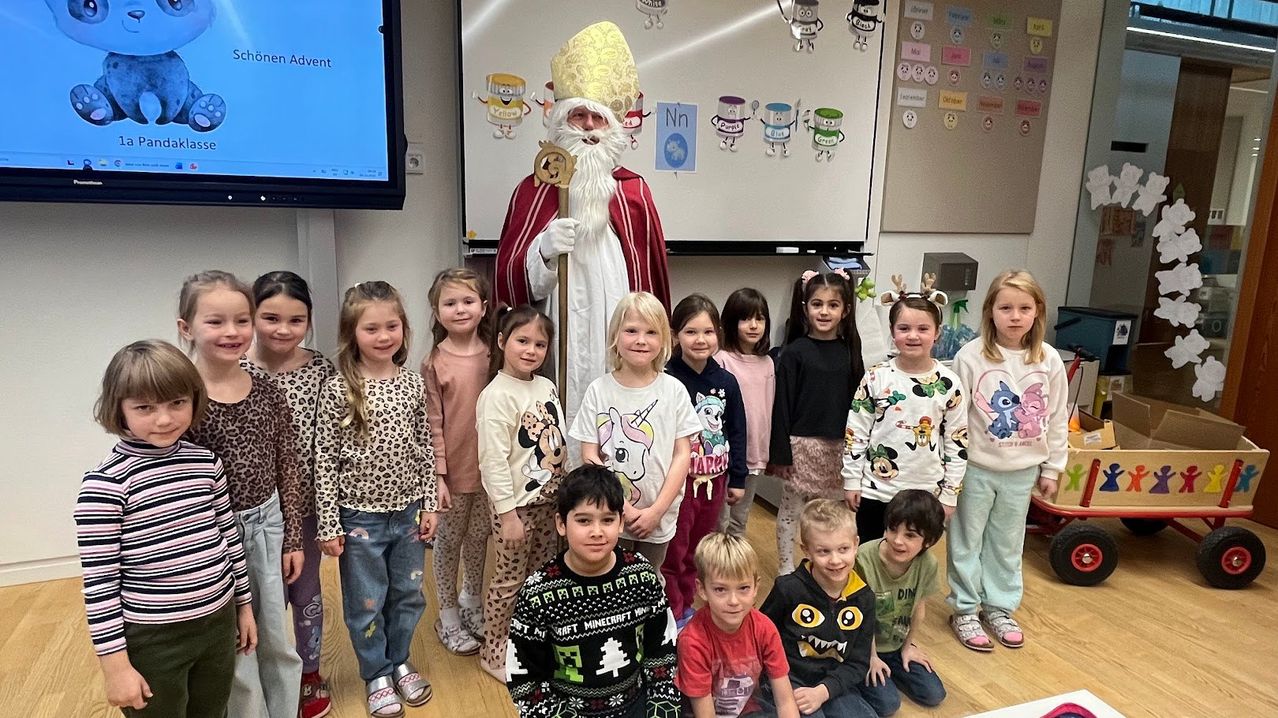 Eine Gruppe von Kindern steht mit einem Mann als Weihnachtsmann verkleidet in einem Klassenzimmer. Dahinter zeigt eine Tafel verschiedene Comicfiguren.