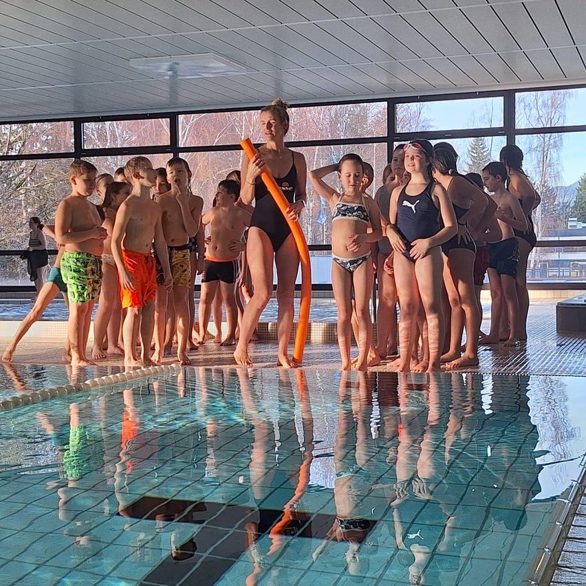 Eine Gruppe von Kindern in Badekleidung steht am Pool, angeführt von einer Frau mit einem Schwimmring. Sie sehen aufmerksam aus, und der Pool reflektiert ihre Bilder.