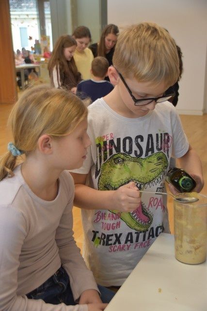 Zwei Kinder sitzen an einem Tisch, ein Junge gießt eine Flasche in einen Becher. Hinter ihnen schauen zwei Mädchen und eine Frau auf einen Tisch.