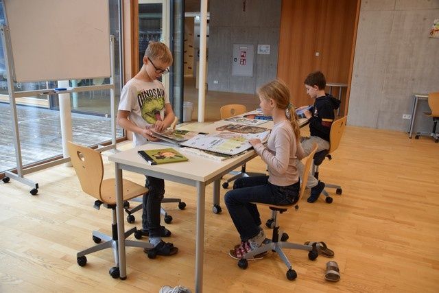 Drei Kinder sitzen an einem Tisch in einem Klassenzimmer, beschäftigt mit Büchern und Papieren. Der Raum ist mit Holzböden und -wänden ausgestattet, mit einer Tafel und Glasfenstern im Hintergrund.