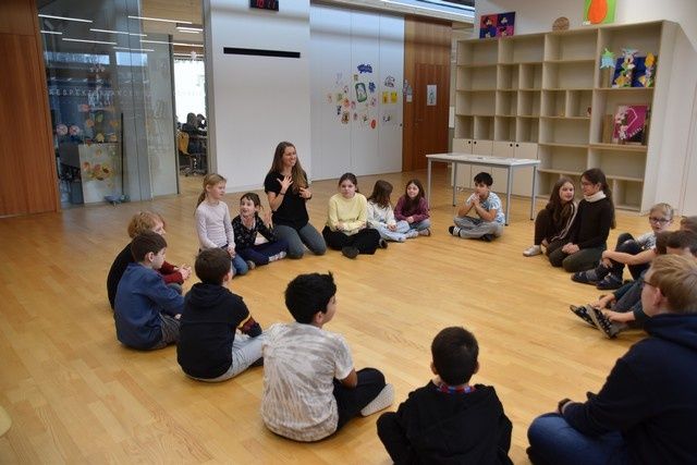 In einem Klassenzimmer sitzen Kinder im Kreis auf dem Boden, während eine Frau in der Mitte steht. Der Raum hat Holzböden, Regale mit Büchern und einen Tisch.