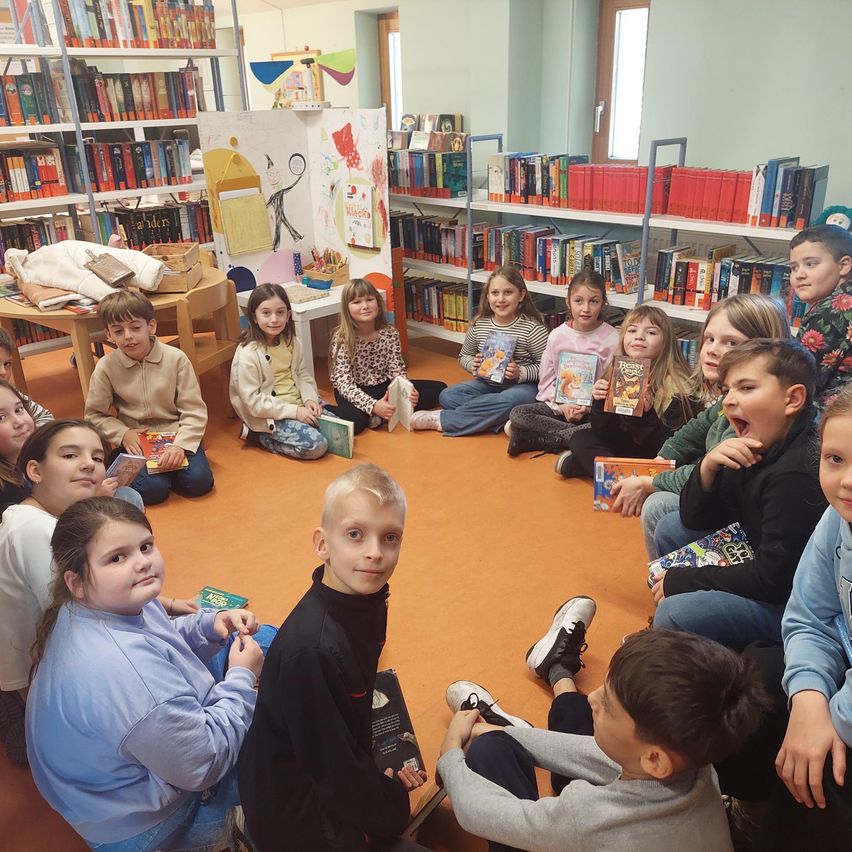 Eine Gruppe von Kindern sitzt in einem Kreis in einer Bibliothek. Sie halten Bücher und scheinen an einer Geschichtensitzung teilzunehmen.