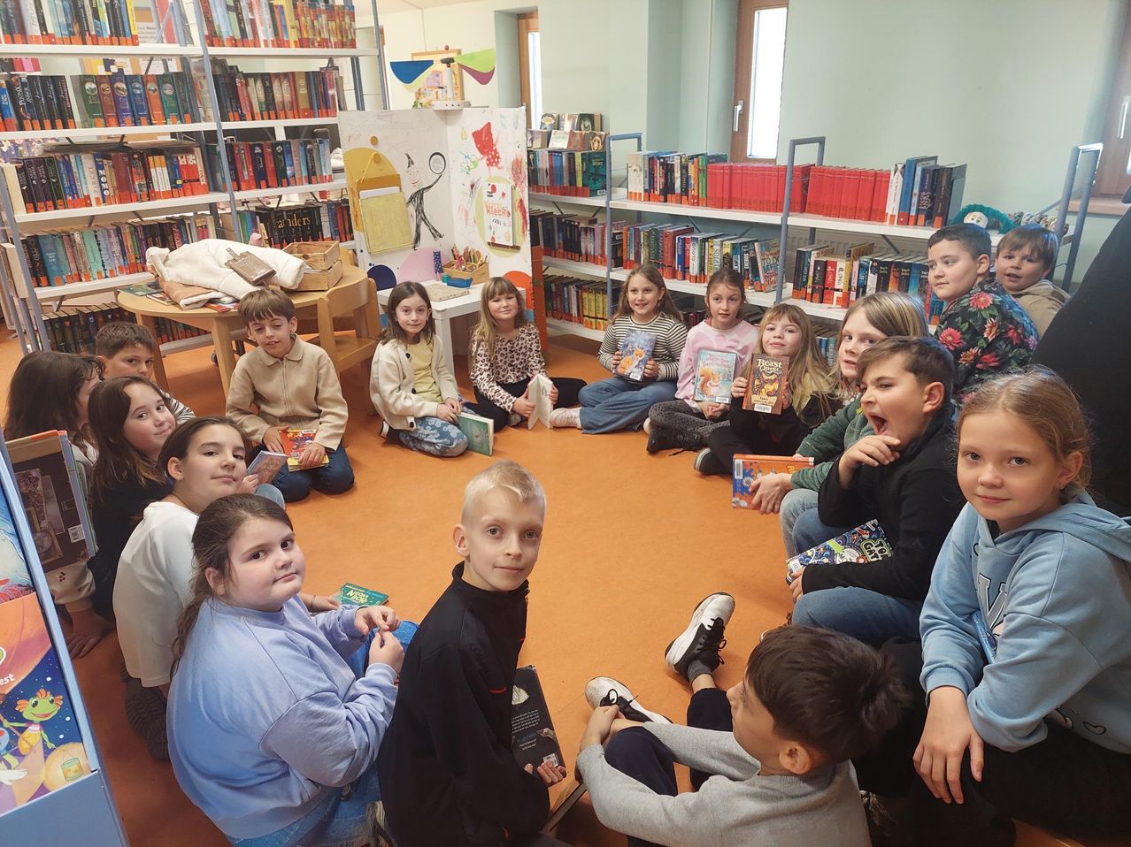 Eine Gruppe von Kindern sitzt in einem Kreis in einer Bibliothek. Sie halten Bücher und scheinen an einer Geschichtensitzung teilzunehmen.