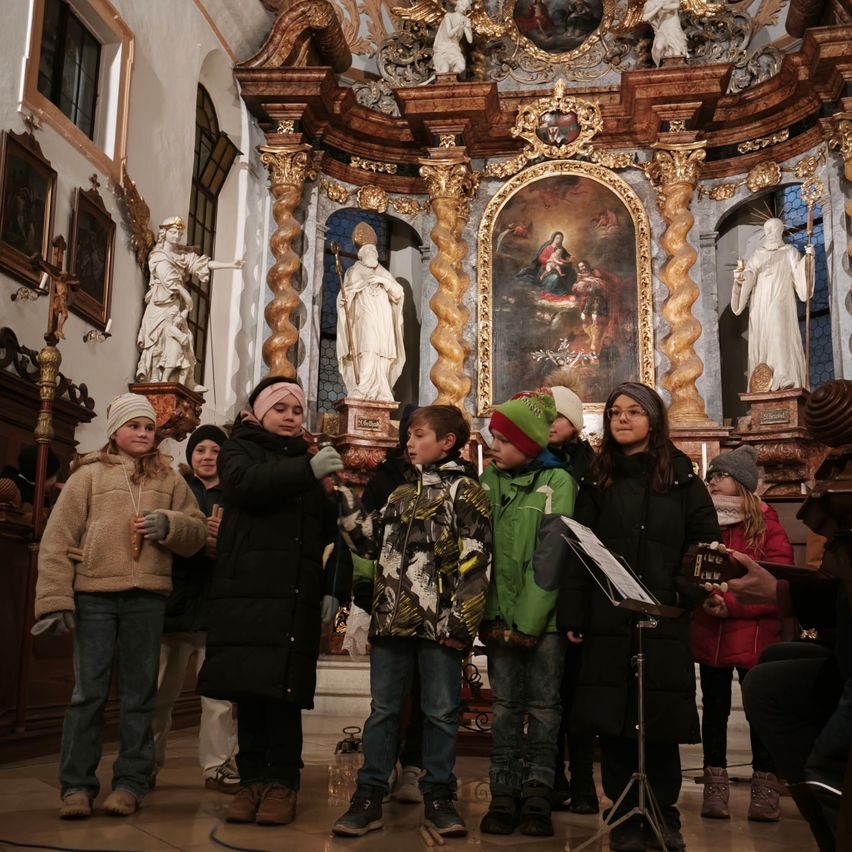 Eine Gruppe von Kindern in Winterkleidung steht in einer Kirche, möglicherweise singen oder beten sie. Hinter ihnen befindet sich ein holzner Altar mit goldenem Design, Statuen und einem Gemälde.