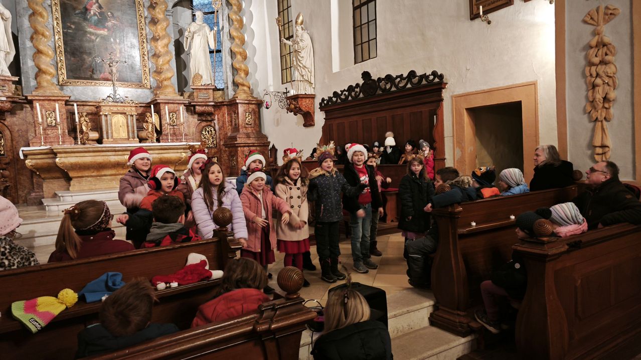 Eine Gruppe von Kindern in Weihnachtsmützen singt in einer Kirche mit Holzbänken und Statuen.