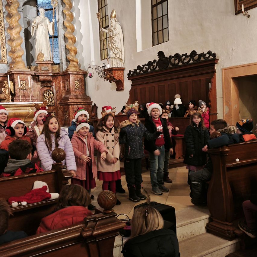 Eine Gruppe von Kindern in Weihnachtsmützen singt in einer Kirche mit Holzbänken und Statuen.