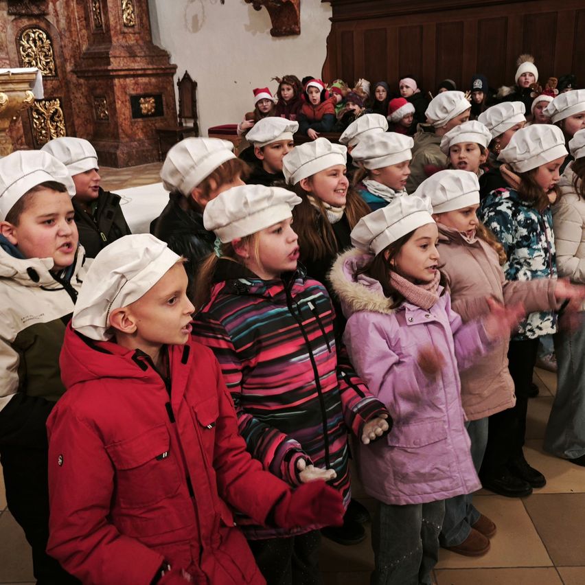 Eine Gruppe von Kindern in Winterkleidung und Kochmützen steht in einer Kirche, möglicherweise singend. Sie schauen nach vorne, und einige halten sich an den Händen. Hinter ihnen stehen weitere Kinder, und einige tragen Weihnachtsmützen. Die Kirche hat Holzwände und einen gefliesten Boden.