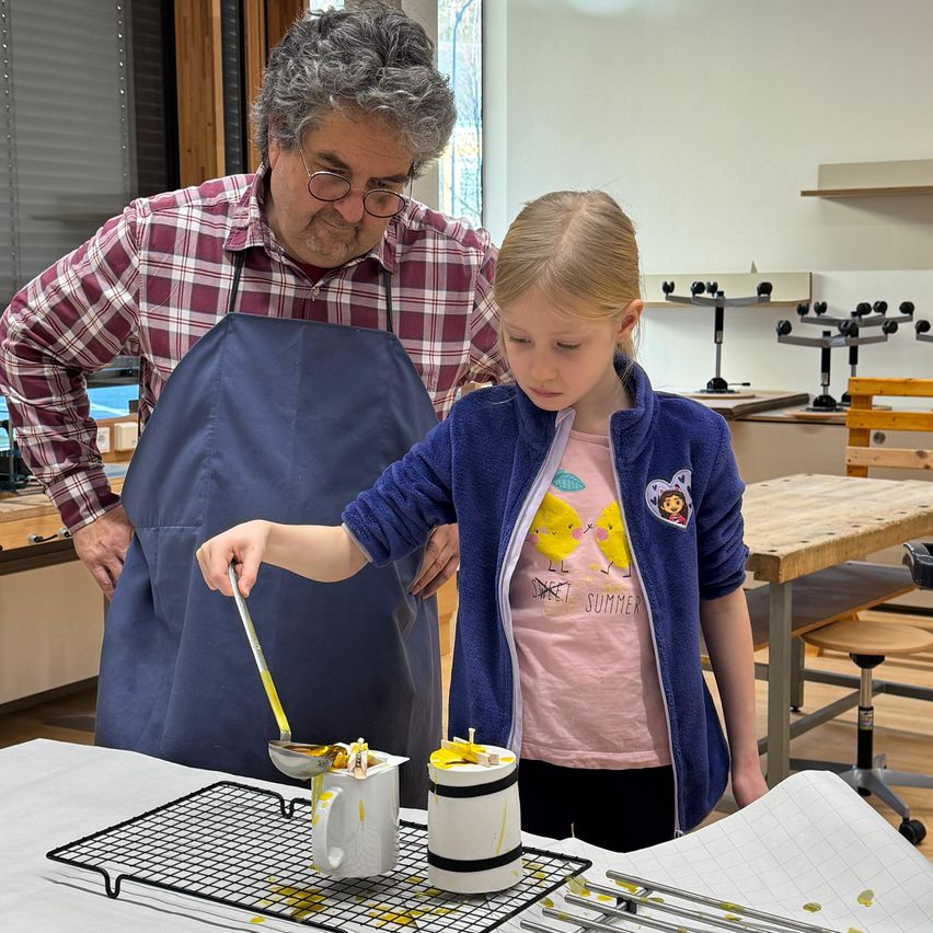 Ein älterer Mann mit Brille und kariertem Hemd steht hinter einem jungen Mädchen in einer blauen Jacke. Sie befinden sich beide in einem Raum mit einem Holztisch und Stühlen. Das Mädchen hält einen Löffel und scheint etwas in einen Becher zu gießen.