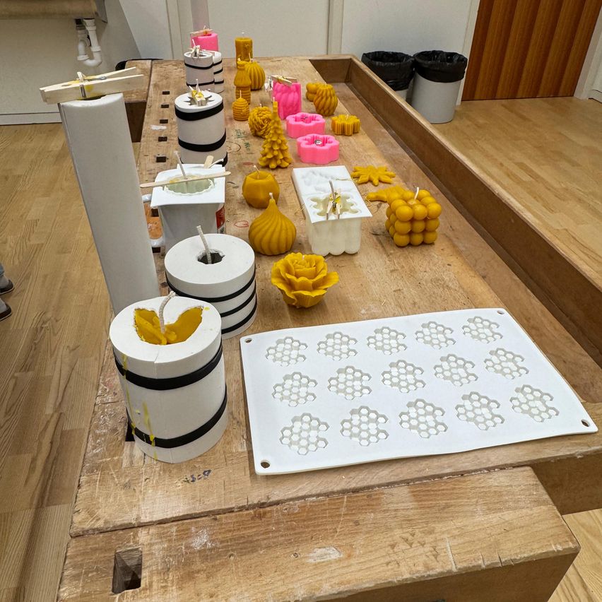 Ein Holztisch zeigt verschiedene Kerzenformen und Kerzen, einschließlich zylindrischer und blumenförmiger Formen, mit einer Honigwabenform auf der rechten Seite. Zwei Mülleimer stehen an der Seite.