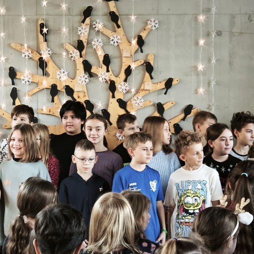 Eine Gruppe von Kindern versammelt sich vor einer Wand, die mit einem holzgeschnitzten Baumdesign, Schneeflocken und hängenden Sternen geschmückt ist, alle festlich gekleidet.