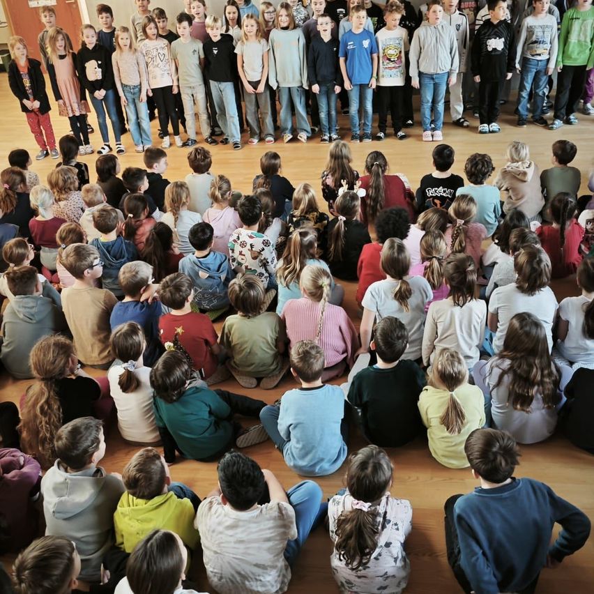 Eine große Gruppe von Kindern und Jugendlichen, einige stehend und einige sitzend, versammelt sich in einem Kreis auf einem Holzboden, möglicherweise für eine Schulveranstaltung.