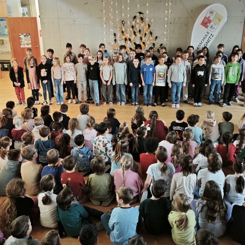 Eine Gruppe von Kindern steht auf der Bühne und singt, während viele weitere Kinder im Kreis um sie herum sitzen. Die Bühne hat einen dekorativen Baum und ein Banner.