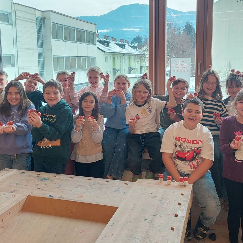 Eine Gruppe von Kindern posiert für ein Foto und hält kleine rote Objekte in die Höhe. Sie stehen vor einem Fenster mit Blick auf einen verschneiten Berg.
