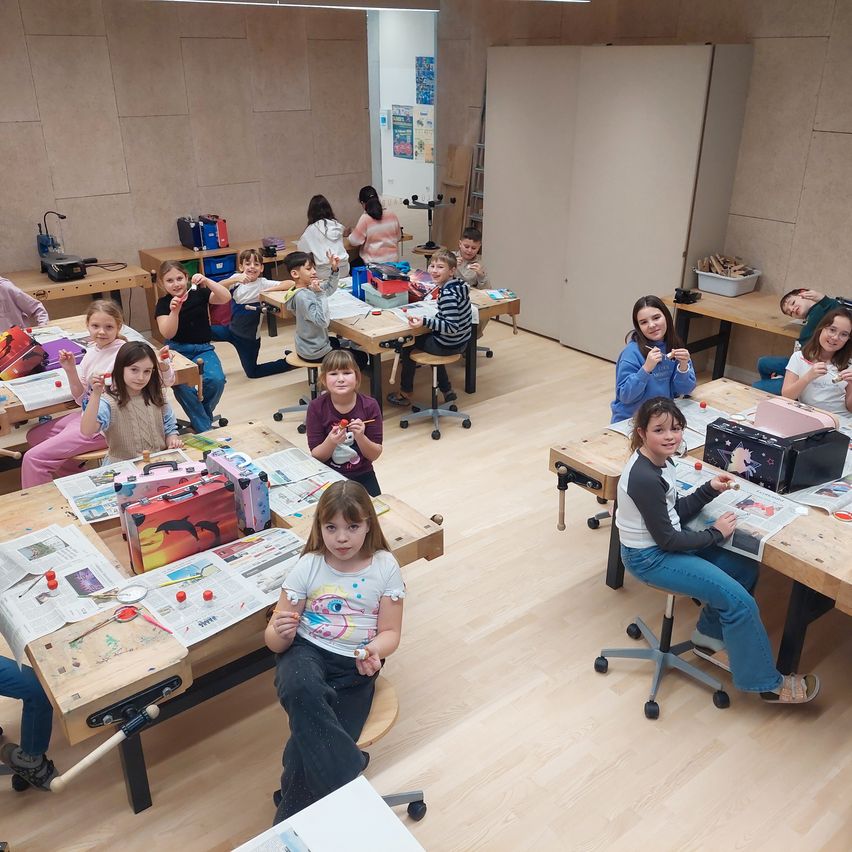 Eine Gruppe von Kindern sitzt in einem Klassenzimmer an Tischen und ist an einem Kunstprojekt beteiligt. Sie verwenden verschiedene Werkzeuge und Materialien. Der Raum hat Holzböden und Wände.