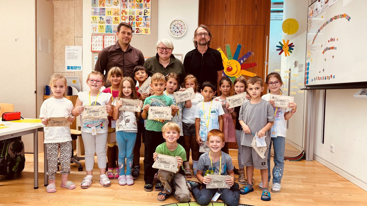 Eine Gruppe von Kindern und drei Erwachsenen steht in einem Klassenzimmer. Sie halten Zertifikate. Zwei Erwachsene stehen hinter den Kindern. Eine Uhr ist an der Wand.