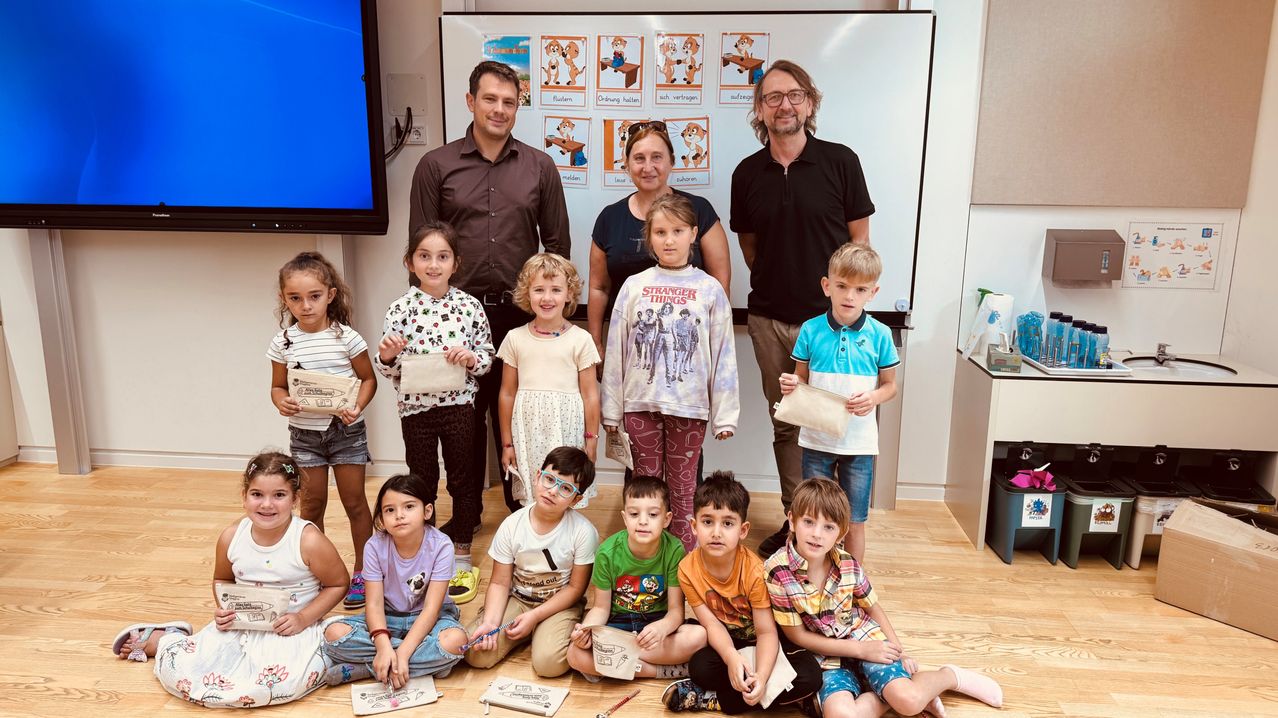 Eine Gruppe von Kindern und Erwachsenen posiert für ein Foto in einem Klassenzimmer. Die Kinder halten Zertifikate. Zwei Erwachsene stehen hinter den Kindern, einer mit Brille. Eine Tafel ist im Hintergrund.
