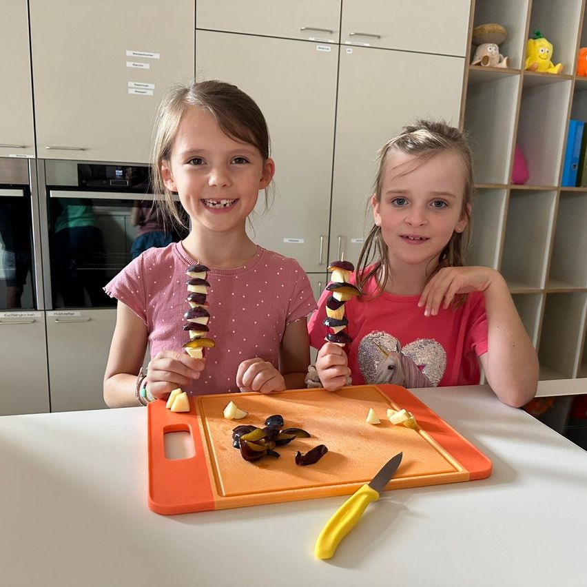 Zwei junge Mädchen bereiten Essen in einer Küche zu, jede hält einen Spieß mit geschnittenen Früchten. Sie sitzen an einem Tisch mit einem Schneidebrett und einem Messer. Hinter ihnen ist eine Küche mit verschiedenen Küchenutensilien und Geräten zu sehen.